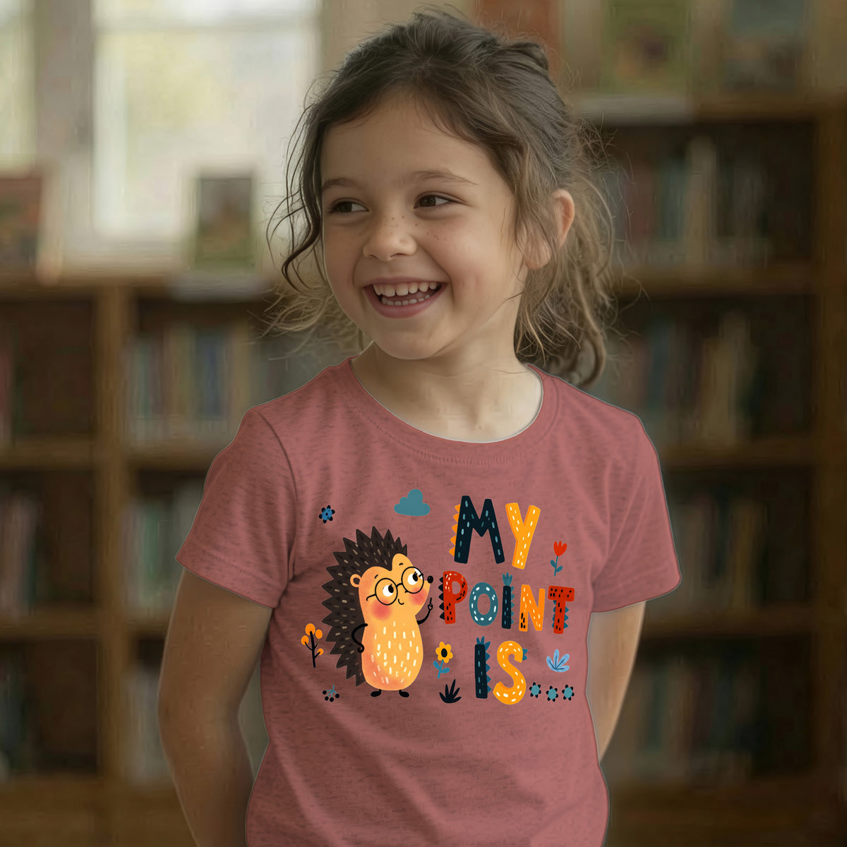 A young girl with brown hair smiling in a library, wearing a heather mauve t-shirt with a colorful graphic of a hedgehog and the text 'MY POINT IS'.