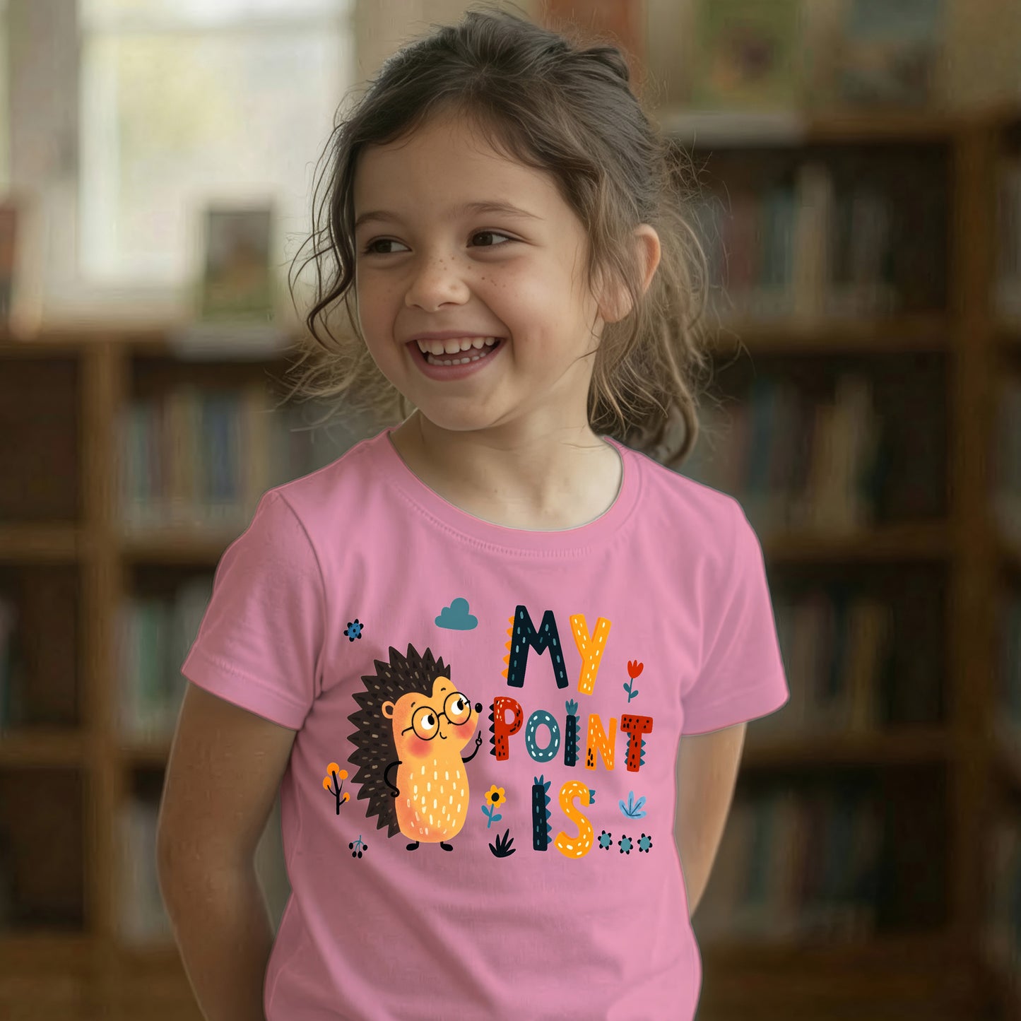 A young girl smiling in a library, wearing a pink t-shirt with a colorful cartoon hedgehog and the text 'MY POINT IS'.