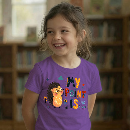 A young girl smiling in a library, wearing a purple t-shirt with a colorful graphic of a hedgehog and the text 'MY POINT IS'.